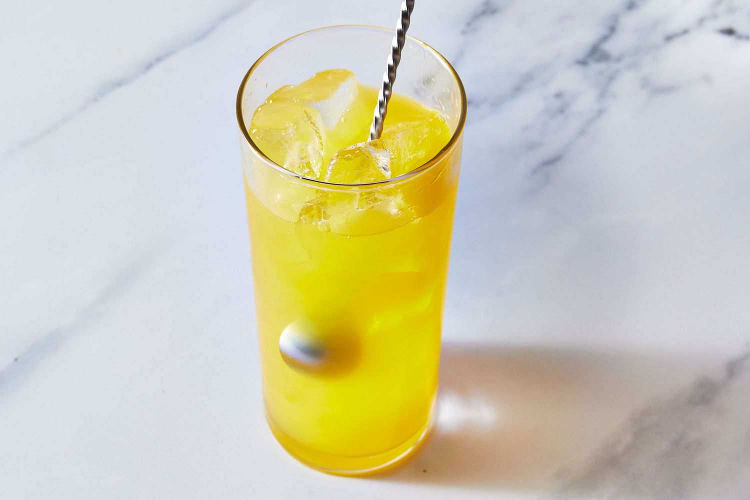 A hand stirring a fuzzy navel cocktail in a tall glass with ice using a bar spoon on a neutral kitchen counter