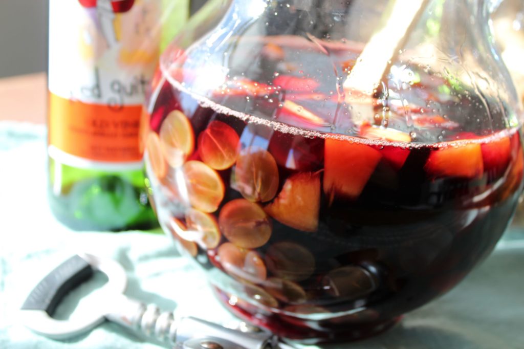 A hand stirring a glass pitcher of red sangria with a long spoon, fruit floating at the top on a kitchen counter