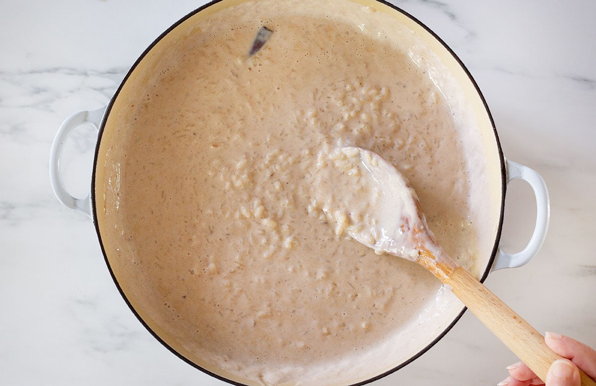A hand stirring creamy arroz con leche in a saucepan with a wooden spoon, cinnamon stick visible in the mixture