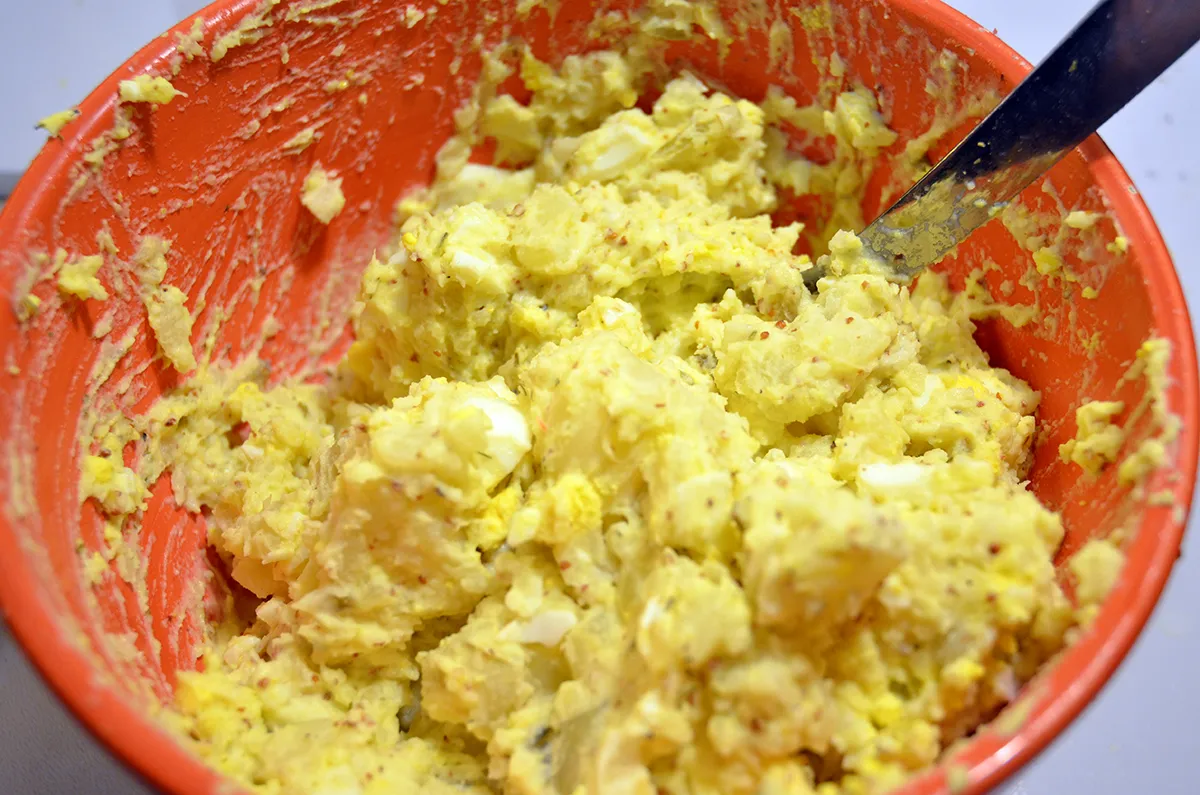 A hand stirring creamy potato salad with a wooden spoon in a large mixing bowl on a kitchen counter
