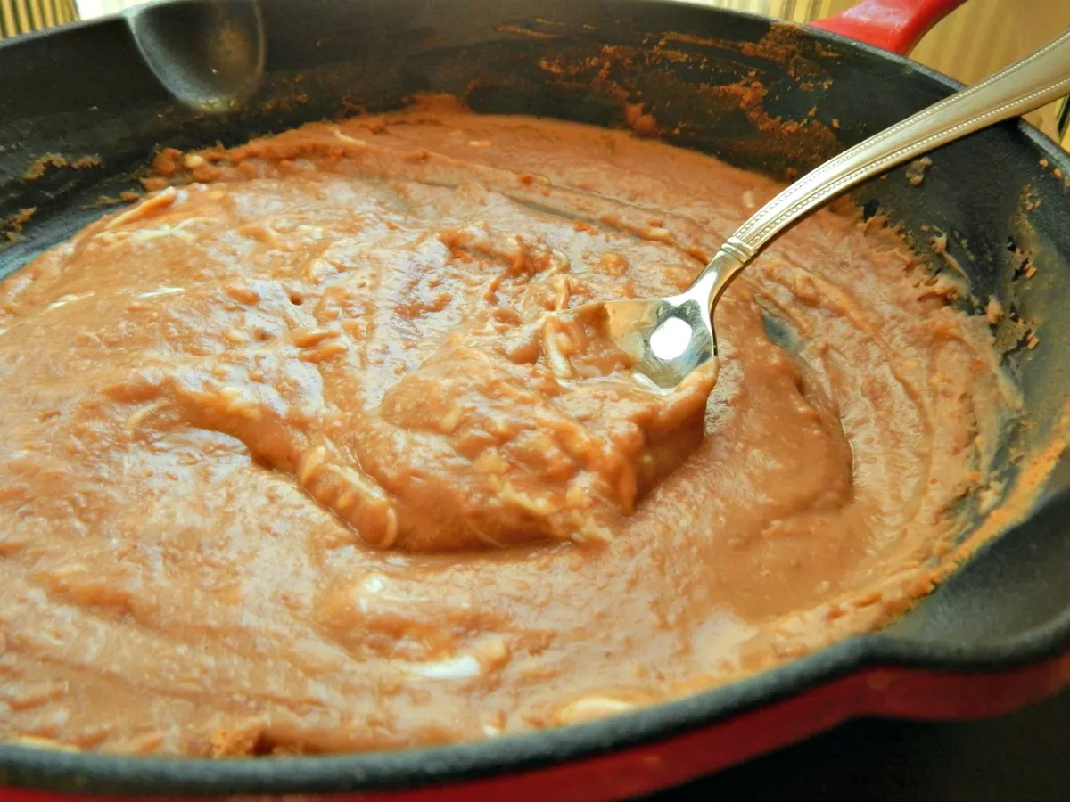 A hand stirring creamy refried beans in a small saucepan on a stovetop with a wooden spoon