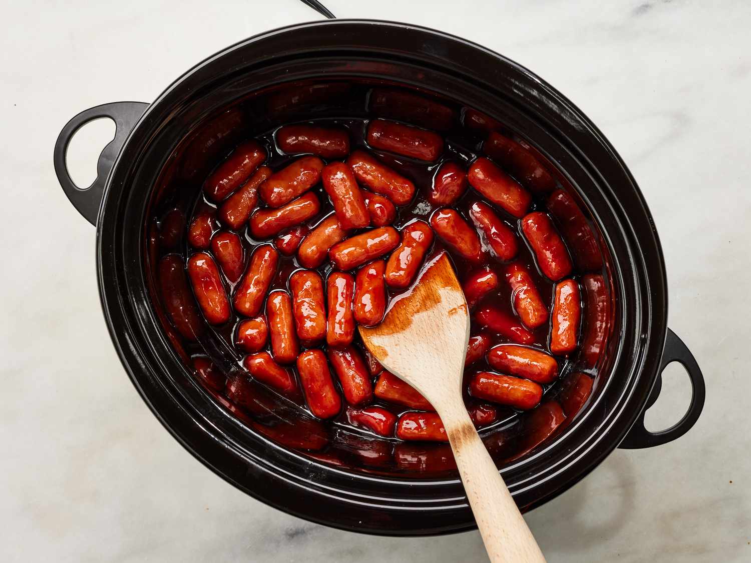 A hand stirring little smokies in a slow cooker as the grape jelly melts into the barbecue sauce, creating a glossy red-brown glaze