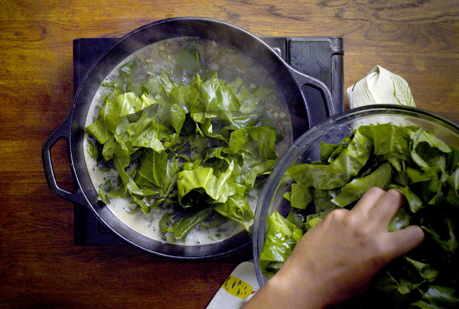 A hand stirring mustard greens in a Dutch oven on the stovetop with steam rising