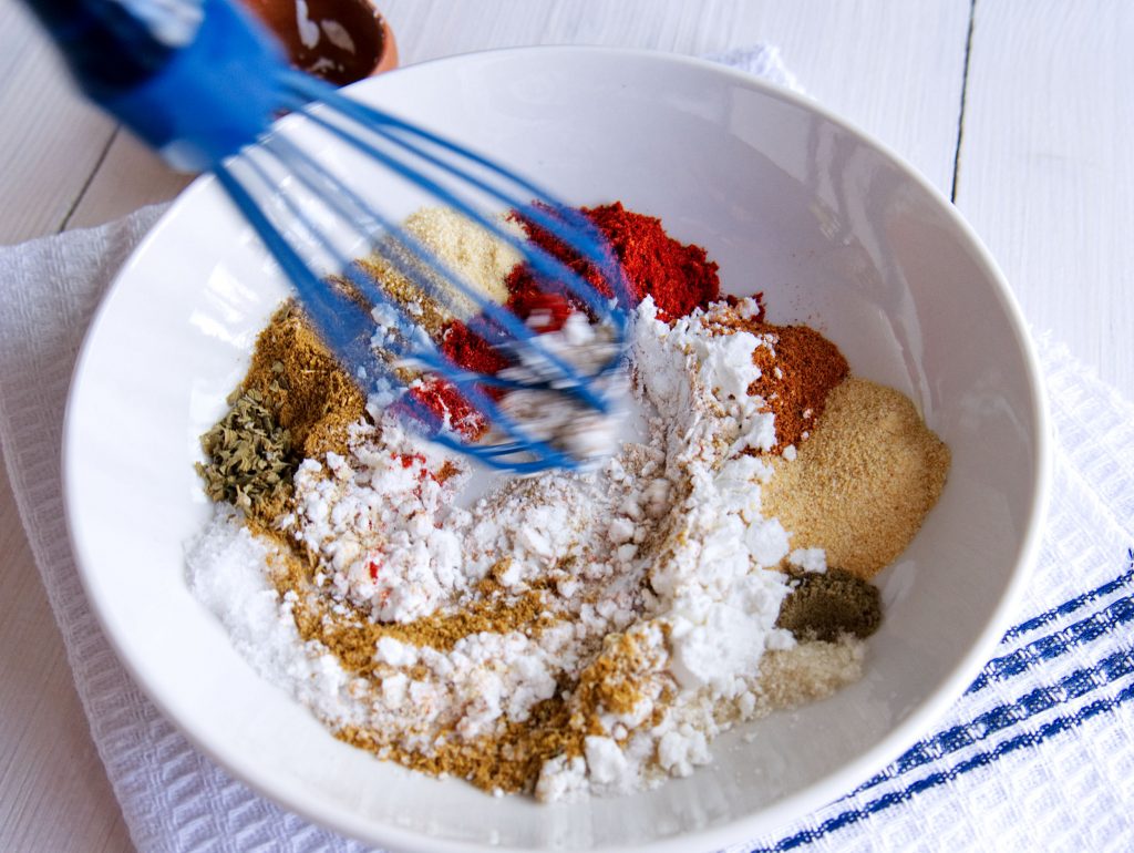 A hand stirring taco seasoning spices together in a small bowl with a spoon on a warm kitchen counter