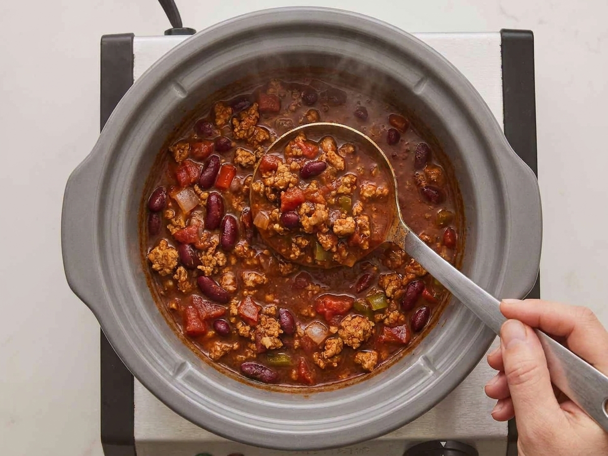 A hand stirring turkey chili inside a slow cooker with a ladle, showing thick beans and tomato base