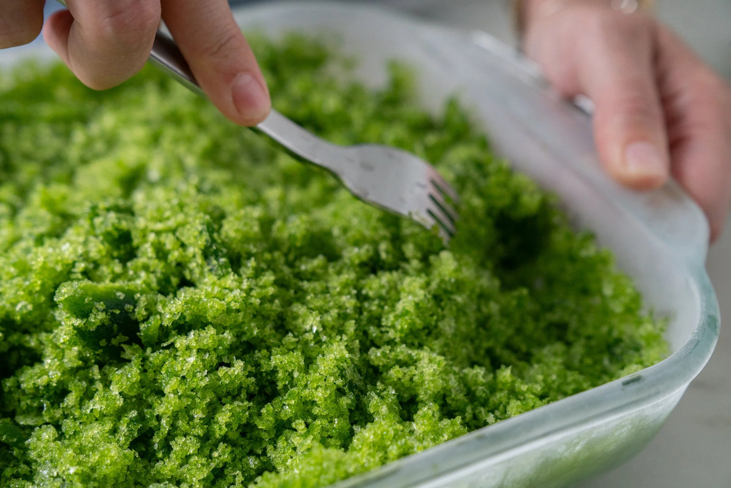 A hand using a fork to scrape icy lemon granita crystals in a shallow metal pan on a kitchen counter