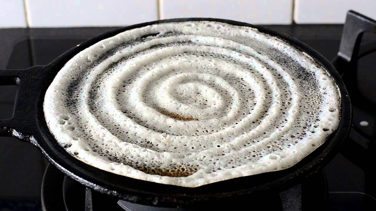 A hand using a ladle to spread dosa batter in quick circles on a hot flat skillet, with steam rising in a home kitchen