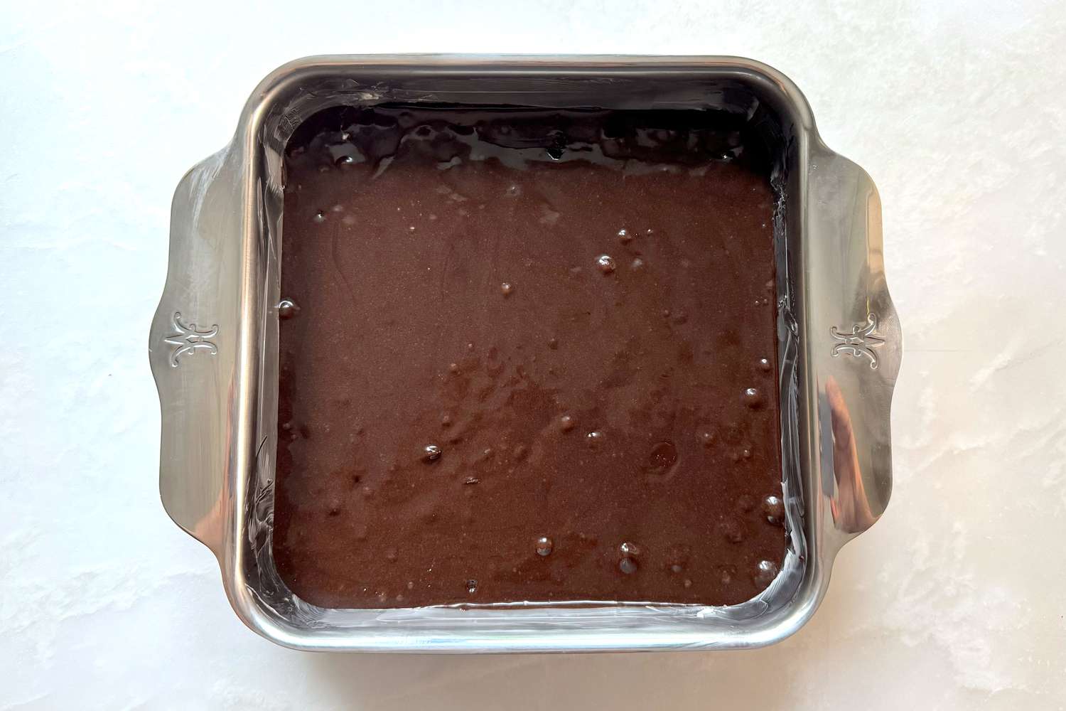 A hand using a rubber spatula to spread glossy brownie batter evenly in a parchment-lined square baking pan