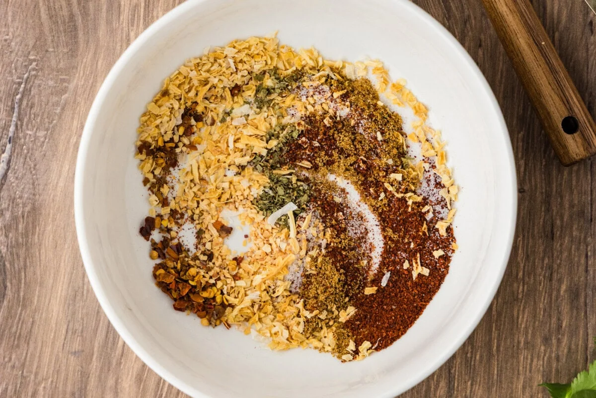 A hand using a small whisk to mix taco seasoning spices in a white ceramic bowl on a kitchen counter