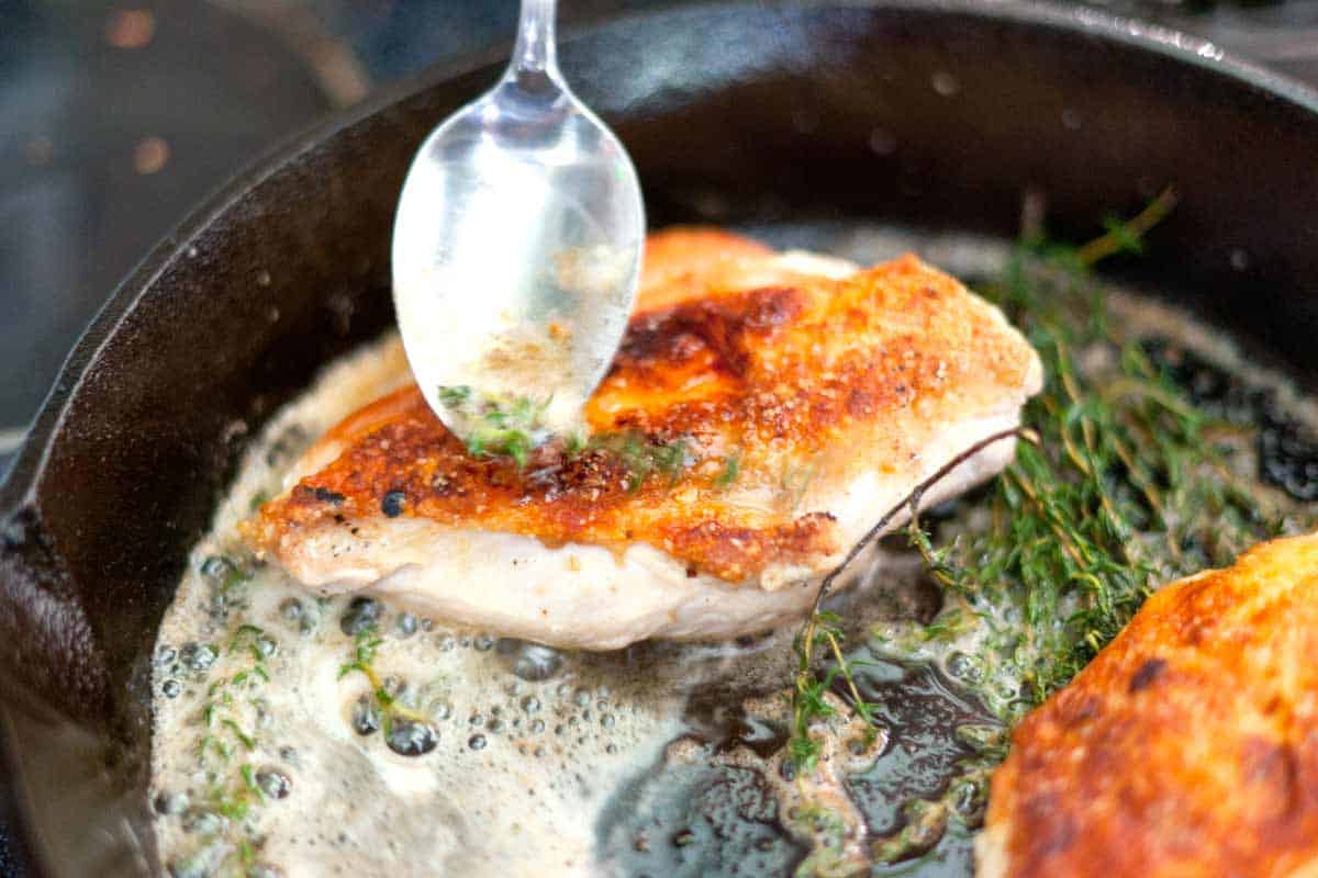 A hand using a spoon to baste a golden roast chicken in a cast iron skillet with bubbling pan juices under warm kitchen lighting