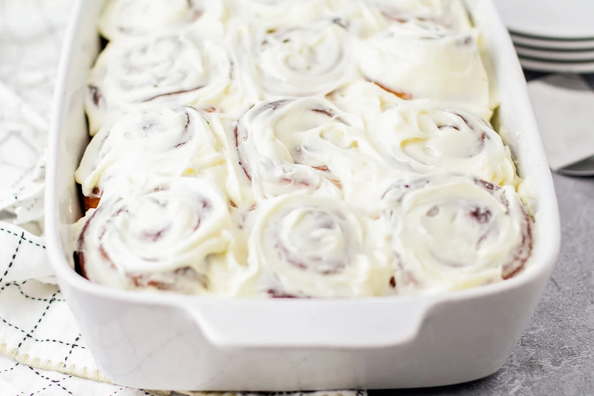 A hand using an offset spatula to spread vanilla frosting over a pan of warm cinnamon rolls