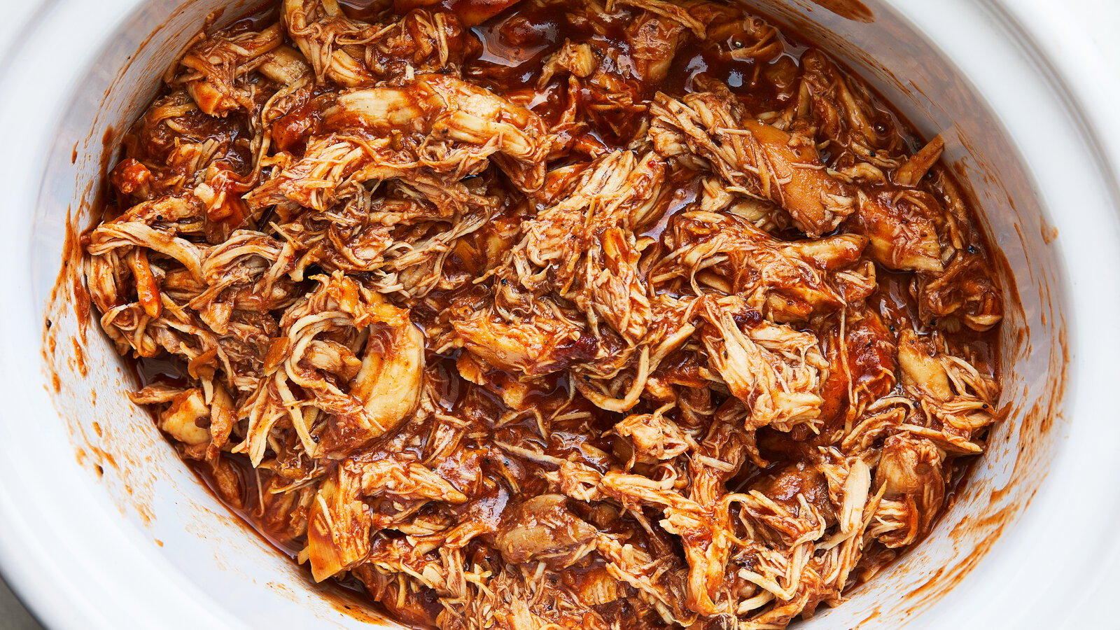 A hand using two forks to shred tender chicken directly in a slow cooker filled with green salsa verde sauce