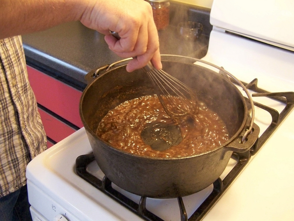 A hand whisking a dark roux in a Dutch oven on a stovetop, with the roux bubbling and turning glossy brown