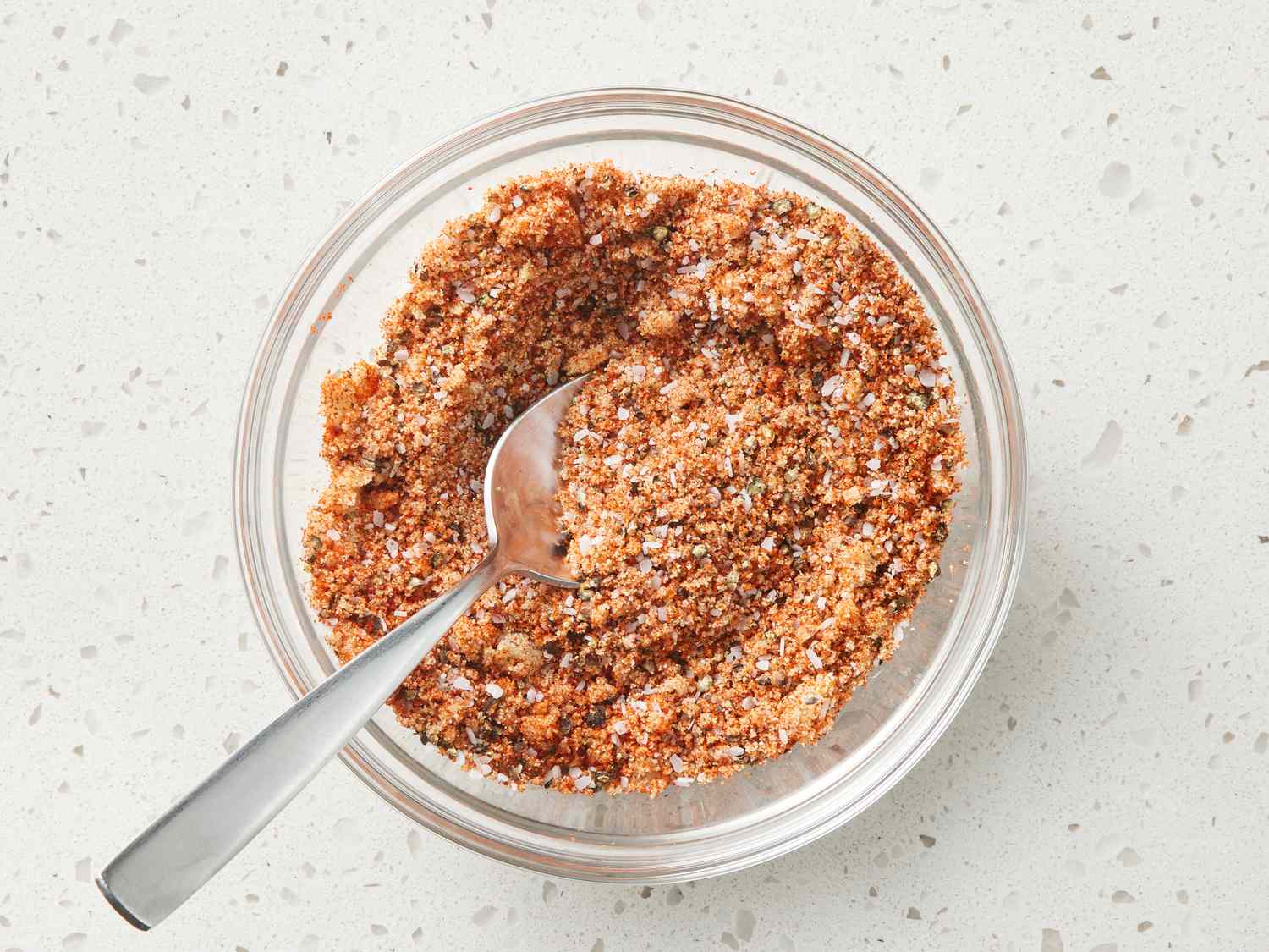 A hand whisking a reddish-brown spice mixture in a small ceramic bowl on a stone countertop