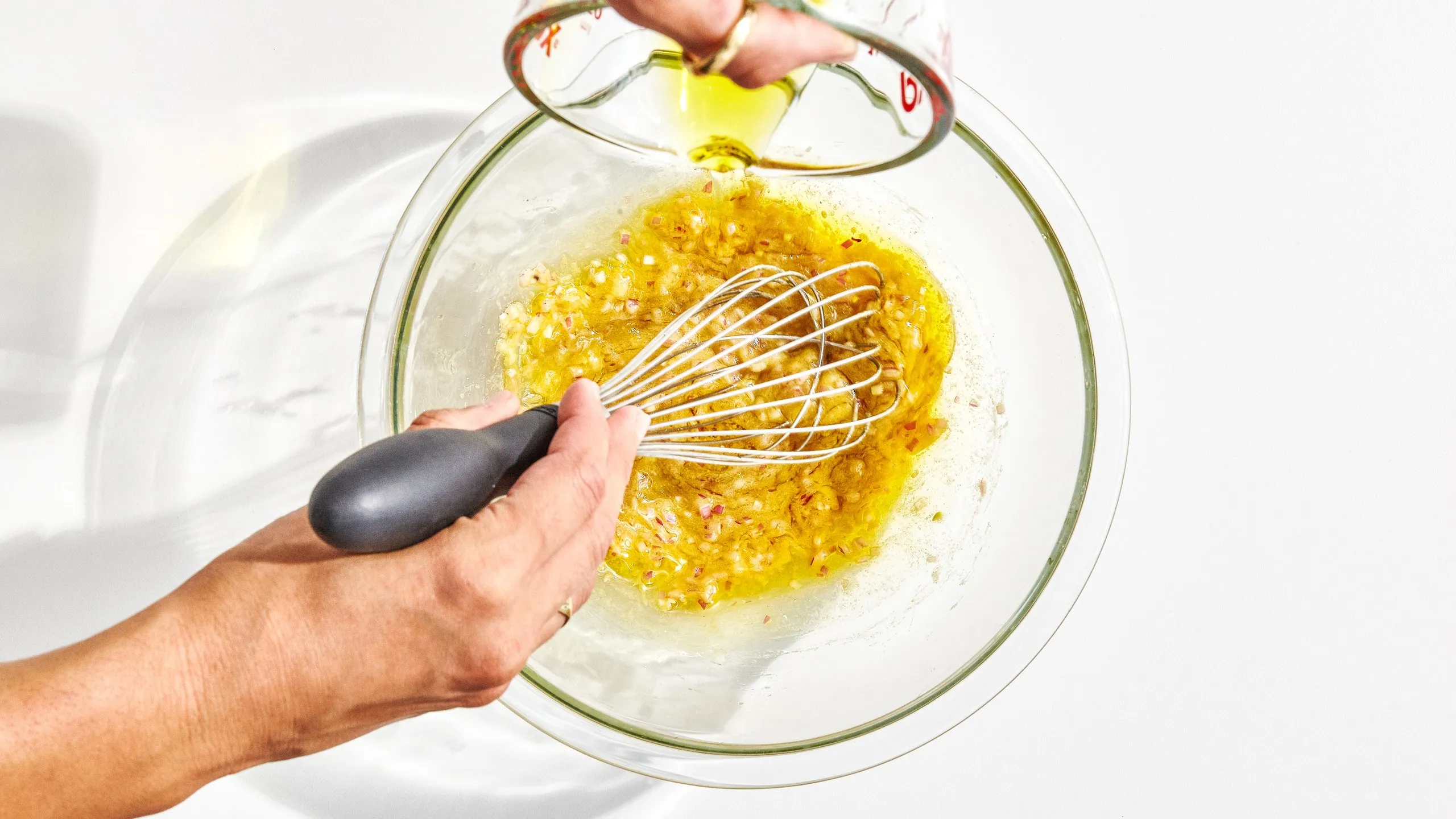 A hand whisking champagne vinaigrette in a small ceramic bowl on a kitchen counter, with olive oil being poured in a thin stream