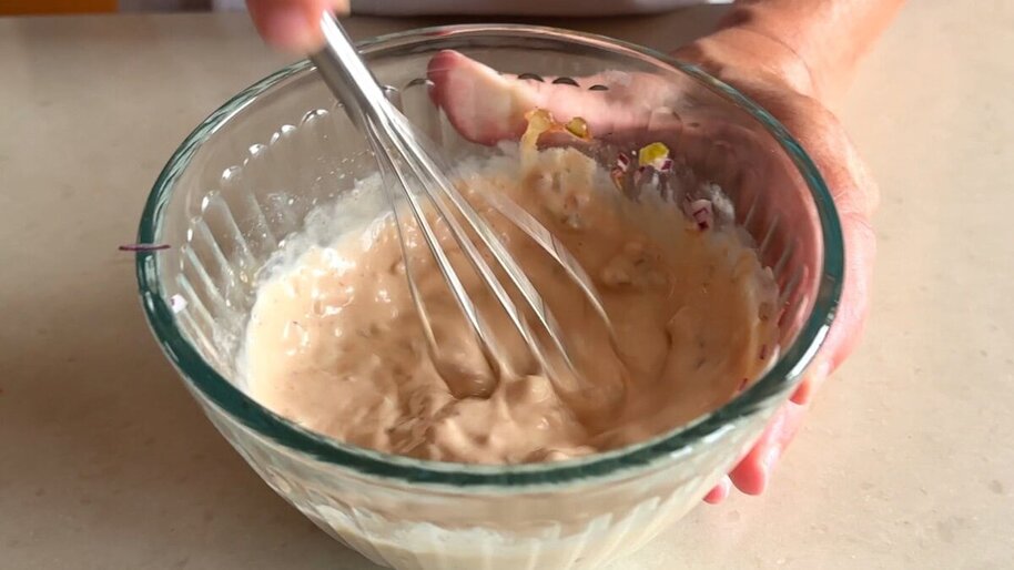 A hand whisking creamy Thousand Island dressing in a glass mixing bowl on a kitchen counter with small ingredient bowls nearby