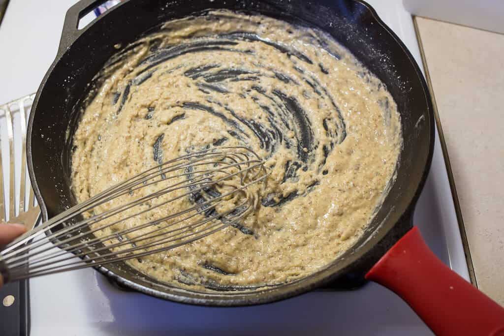 A hand whisking creamy pepper gravy in a cast iron skillet on a stovetop with browned bits dissolving into the sauce