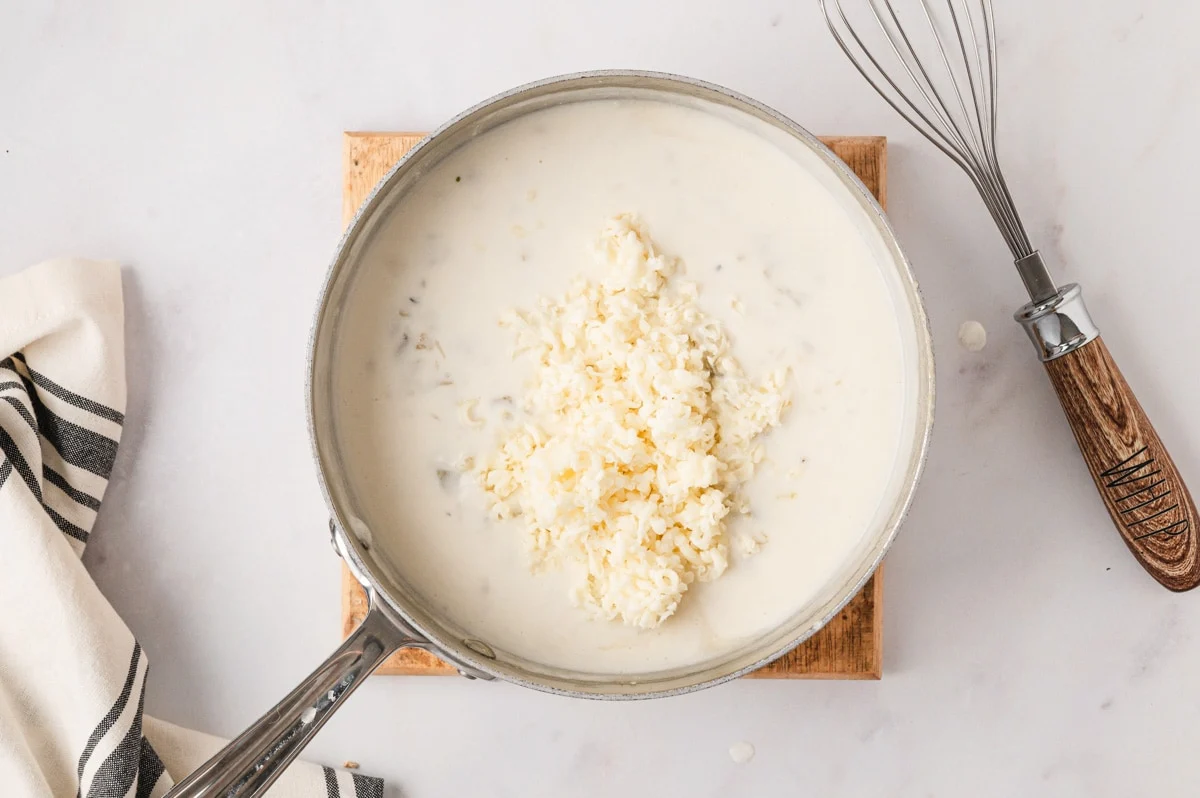 A hand whisking creamy white enchilada sauce in a saucepan on a stovetop, with steam rising gently under warm kitchen lighting