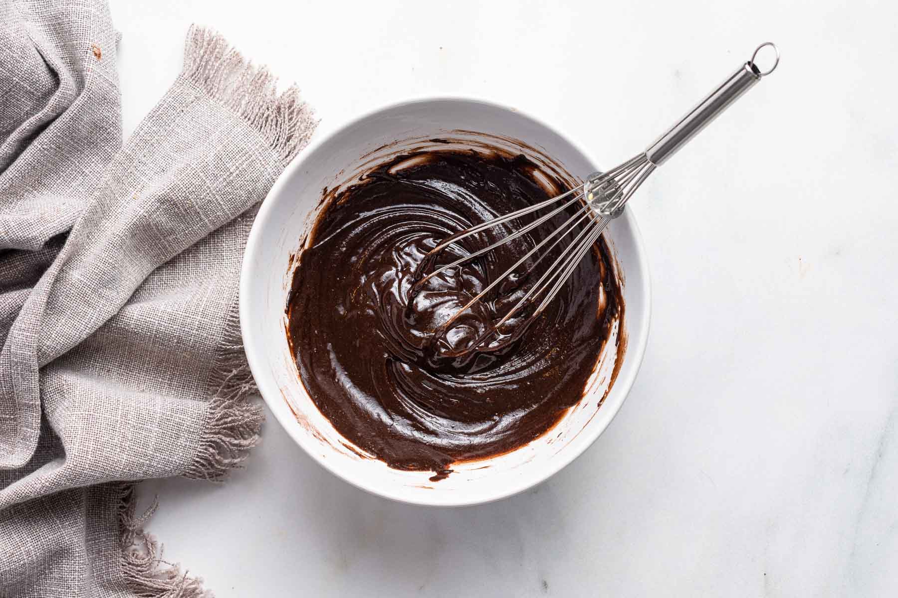 A hand whisking glossy cocoa brownie batter in a large mixing bowl on a kitchen counter