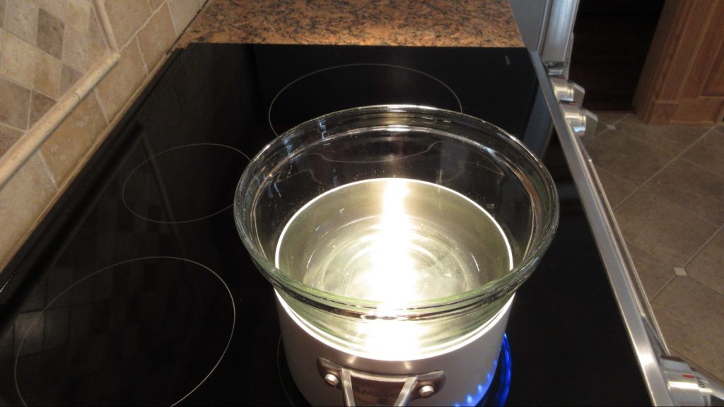 A heatproof bowl set over a saucepan of simmering water on a stovetop, with egg whites and sugar being whisked together for Swiss meringue