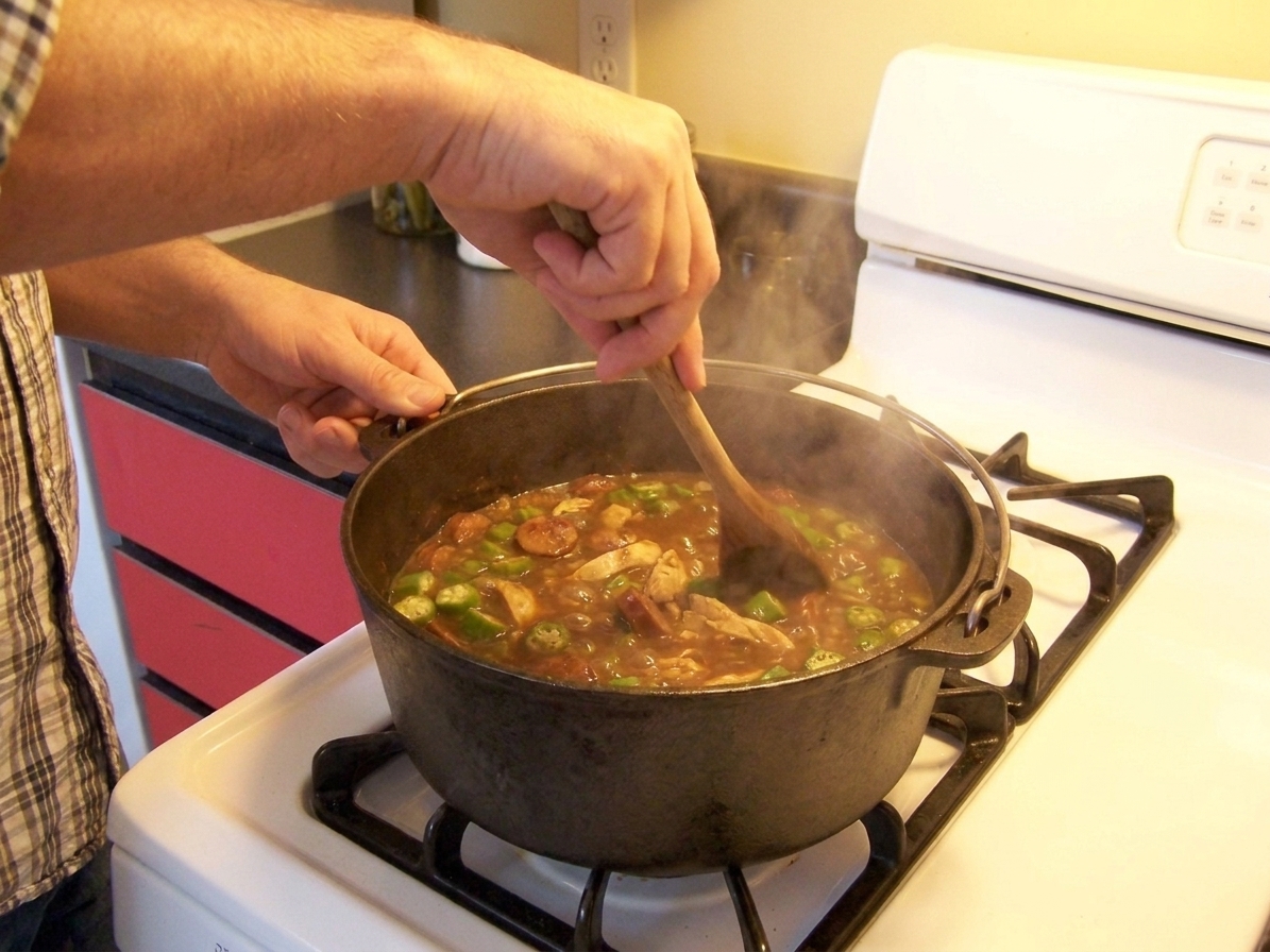A home cook stirring a pot of gumbo on a stovetop with a wooden spoon, with steam rising in a warmly lit kitchen