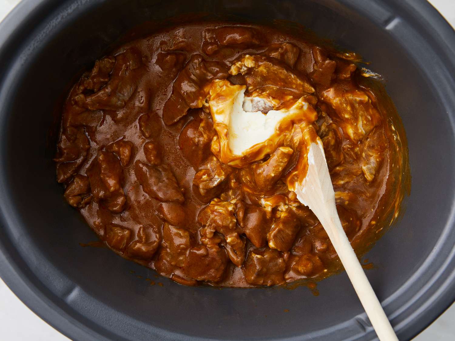 A home cook stirring creamy beef stroganoff in a slow cooker with a wooden spoon on a clean kitchen counter