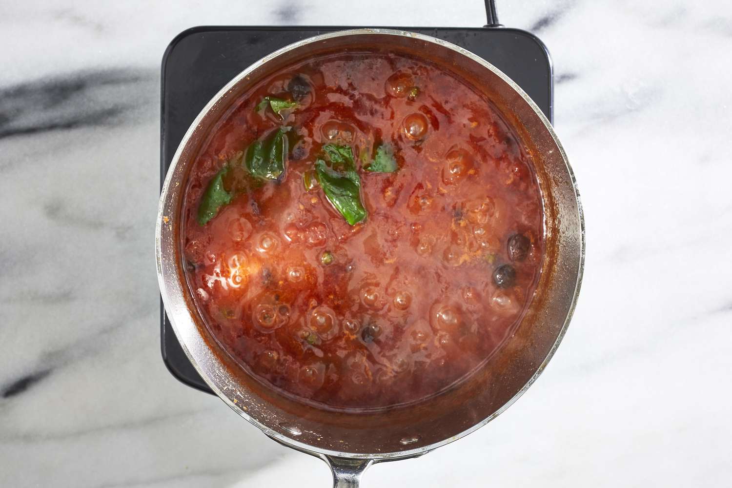 A home cook stirring pasta puttanesca sauce in a wide skillet on a stovetop, with steam rising and a wooden spoon mid-stir, warm kitchen lighting