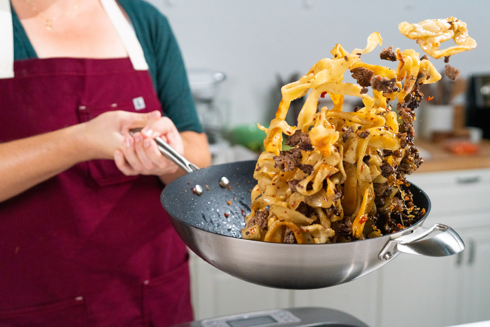 A home cook using tongs to toss noodles in a skillet with glossy sauce on a stovetop under warm kitchen lighting