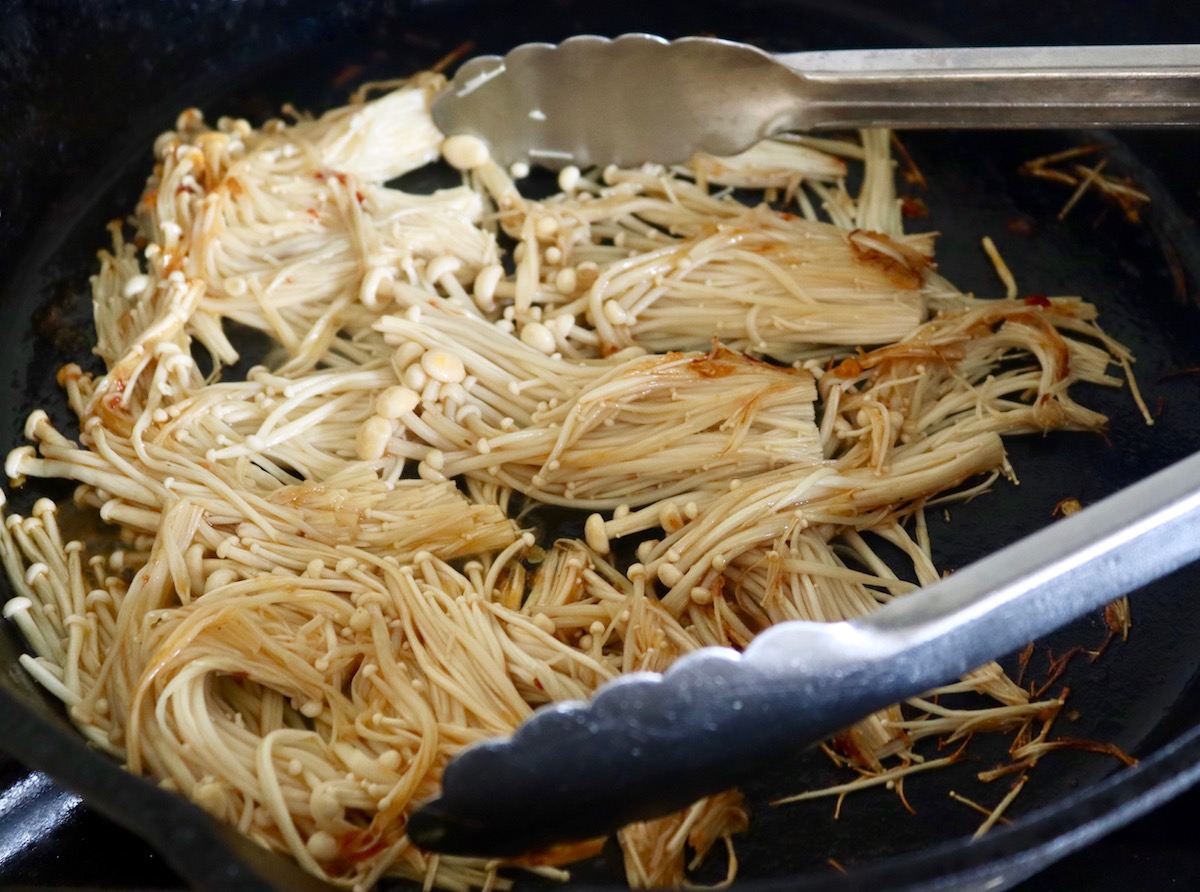 A home cook using tongs to turn browned enoki mushroom bundles in a hot skillet on a stovetop