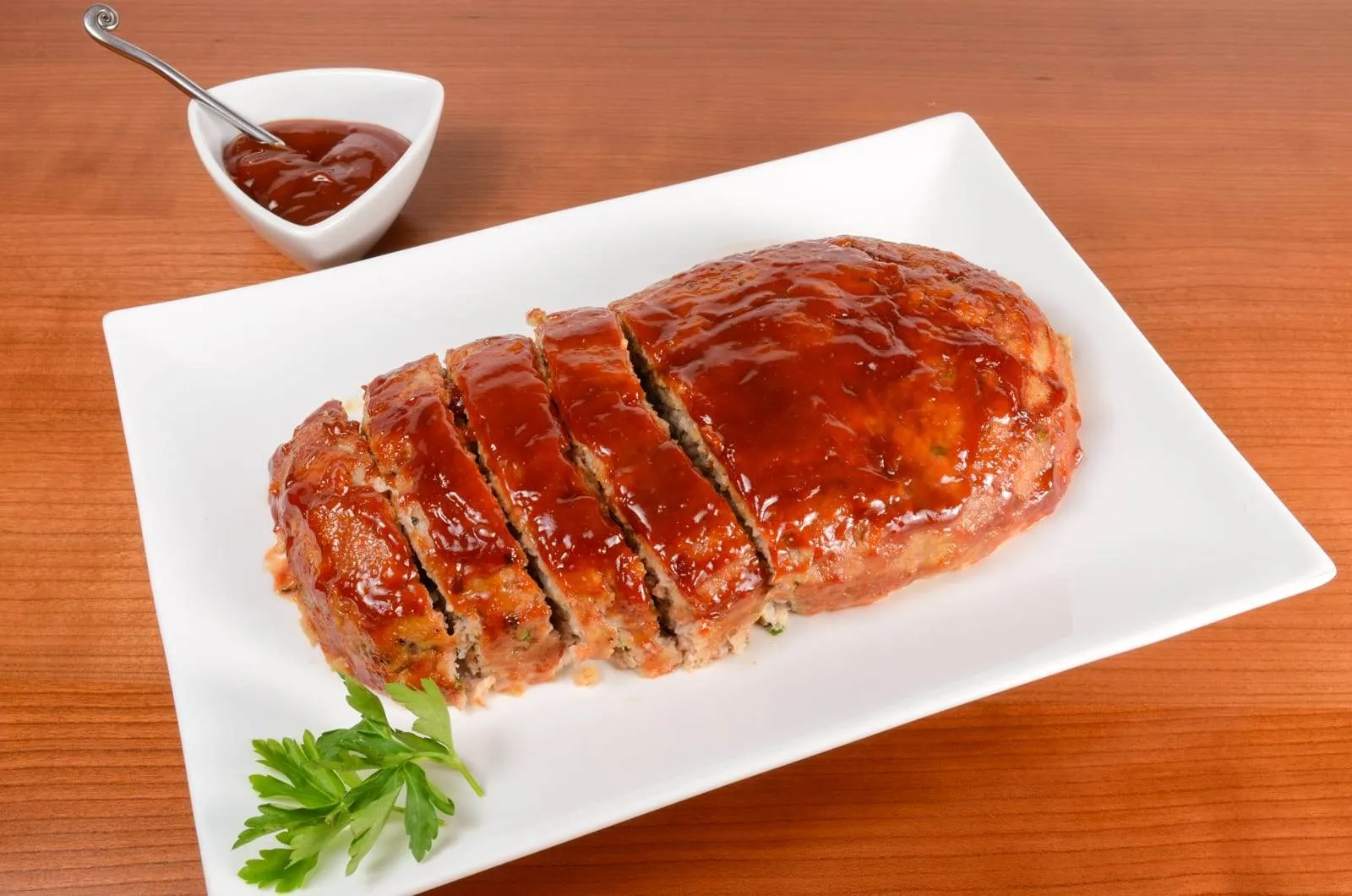 A home cook's hands brushing ketchup glaze over an uncooked meatloaf shaped on a parchment-lined baking sheet in a warm home kitchen