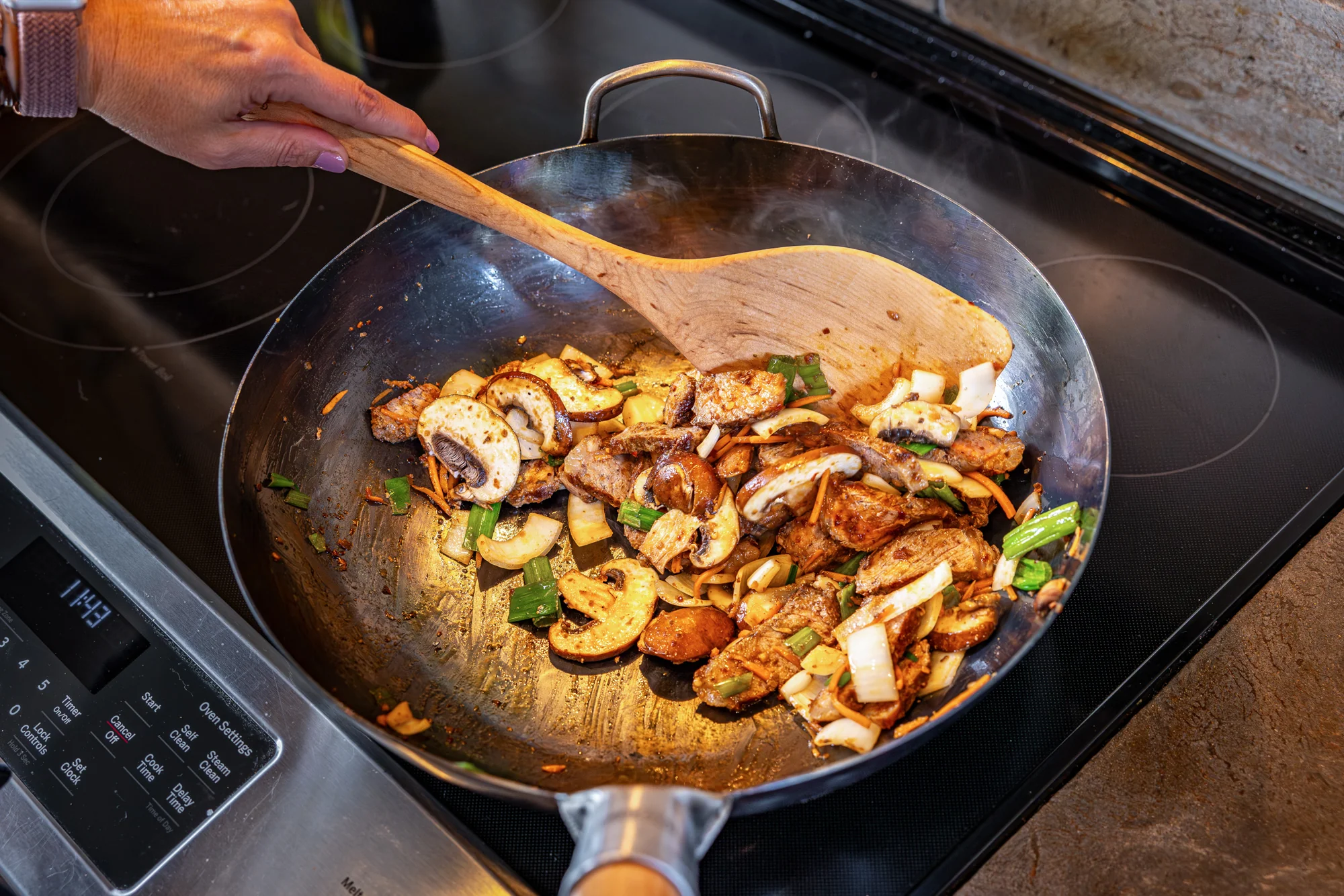 A home cook’s hands holding a wok spatula over a large skillet on a stovetop, ready to stir-fry noodles