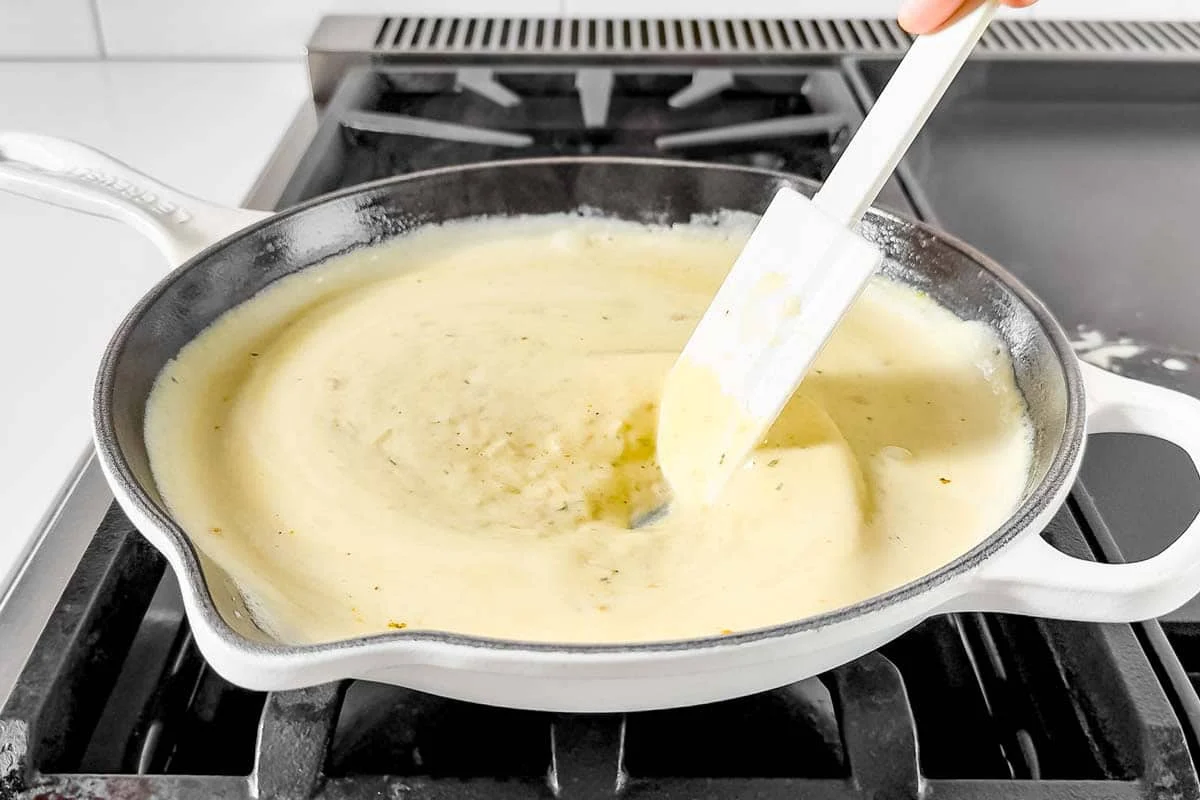 A home cook's hands stirring a creamy Boursin sauce in a skillet on the stovetop with a wooden spoon, warm kitchen lighting