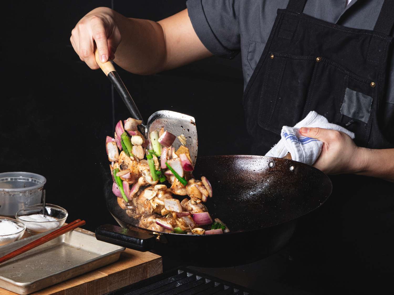 A home cook's hands tossing chicken and vegetables in a hot skillet on a stovetop with a wooden spatula, warm kitchen lighting