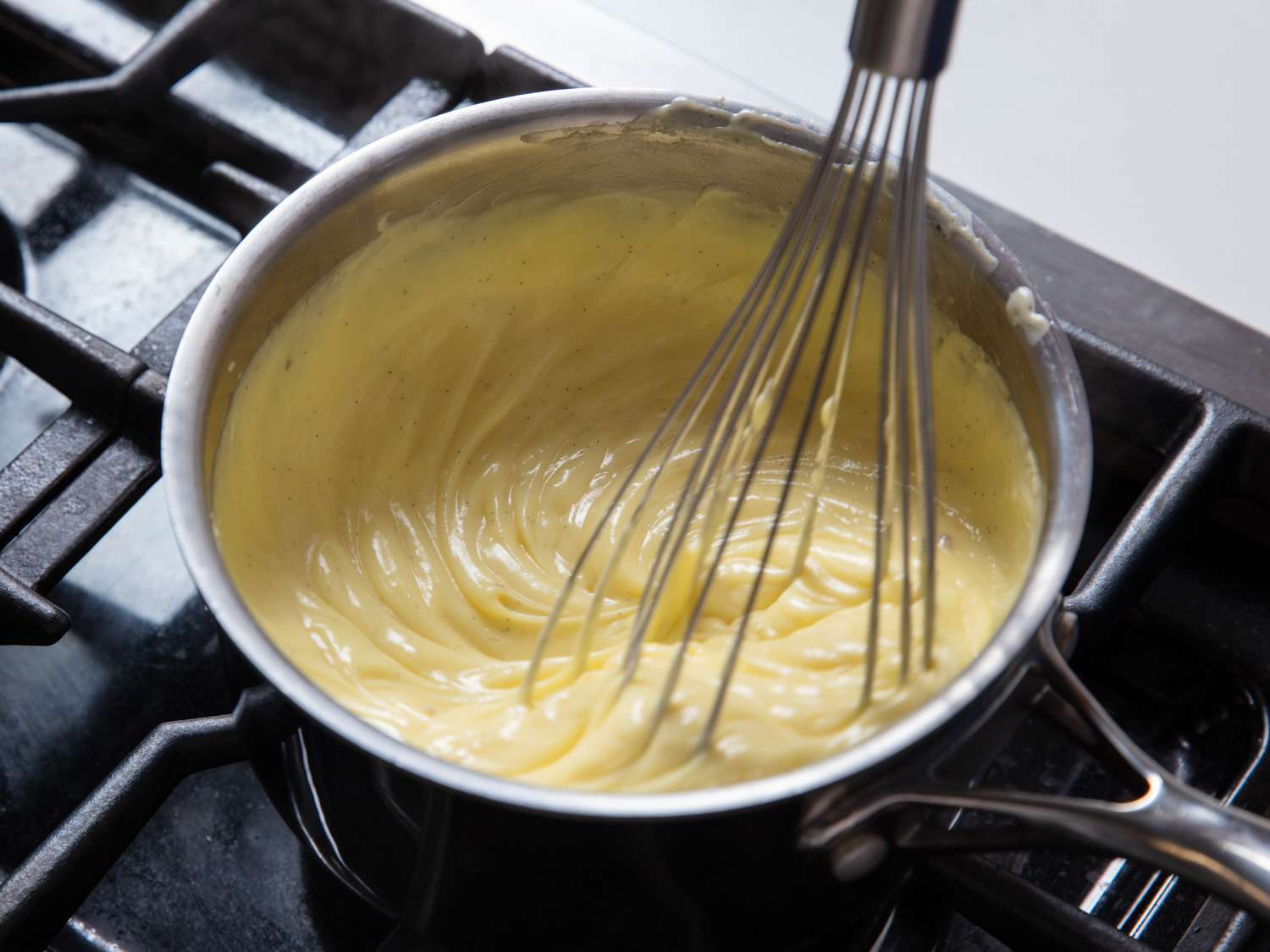 A home cook's hands whisking thick vanilla pastry cream in a saucepan on a stovetop, with a wooden spoon and small bowls nearby on a tidy counter