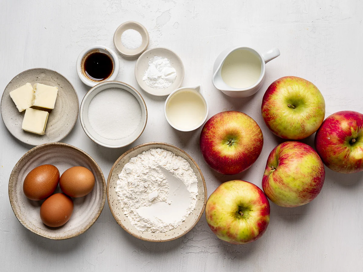 A home kitchen counter with flour, sugar, butter, eggs, vanilla, spices, and two apples laid out for baking