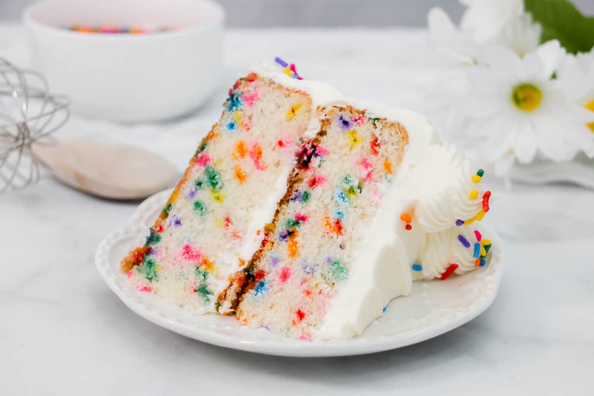 A homemade confetti layer cake with vanilla buttercream, sliced to show a sprinkle-speckled crumb, on a white cake stand on a bright kitchen counter