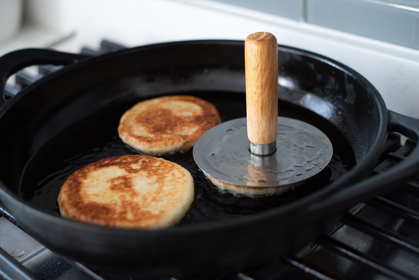 A hotteok pancake frying in a nonstick skillet as it is gently pressed flat with a spatula, with a lid nearby on a clean stovetop
