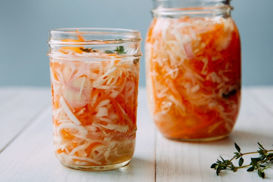 A jar of Haitian pikliz with shredded cabbage, carrots, and peppers in brine sitting on a kitchen counter next to a small bowl