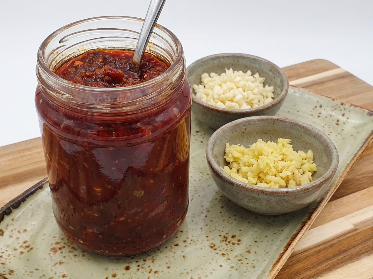 A jar of doubanjiang (fermented chili broad bean paste) sitting on a kitchen counter beside small bowls of minced garlic and ginger