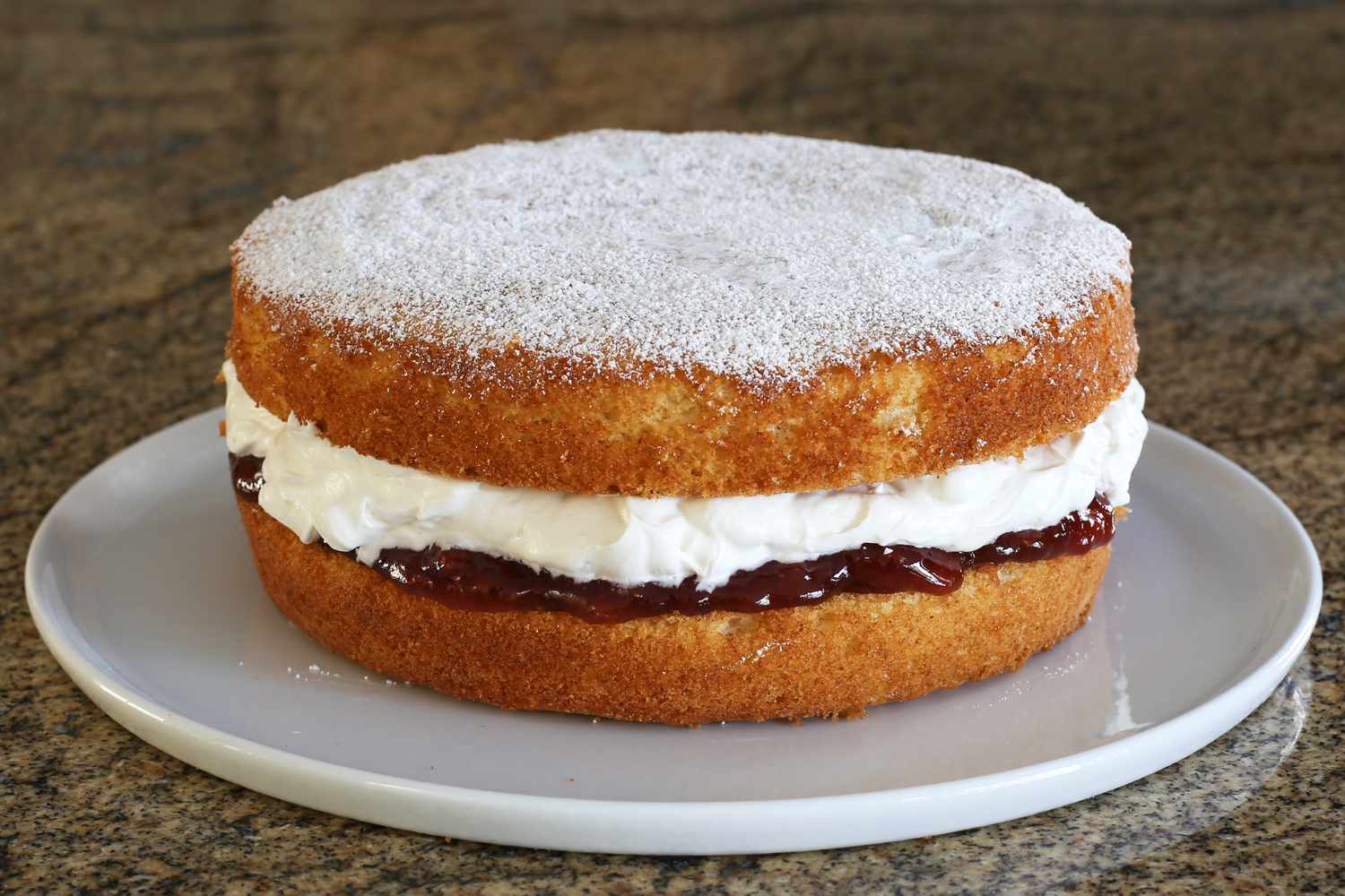 A jar of strawberry jam, a bowl of whipped cream, and a sieve with powdered sugar on a kitchen counter ready for assembling a Victoria sponge cake