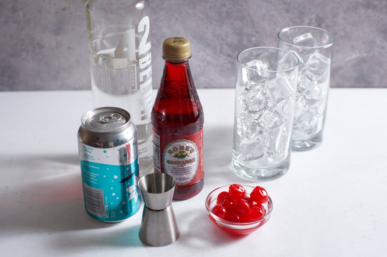A jigger of grenadine on a kitchen counter next to ginger ale, ice, and cherries