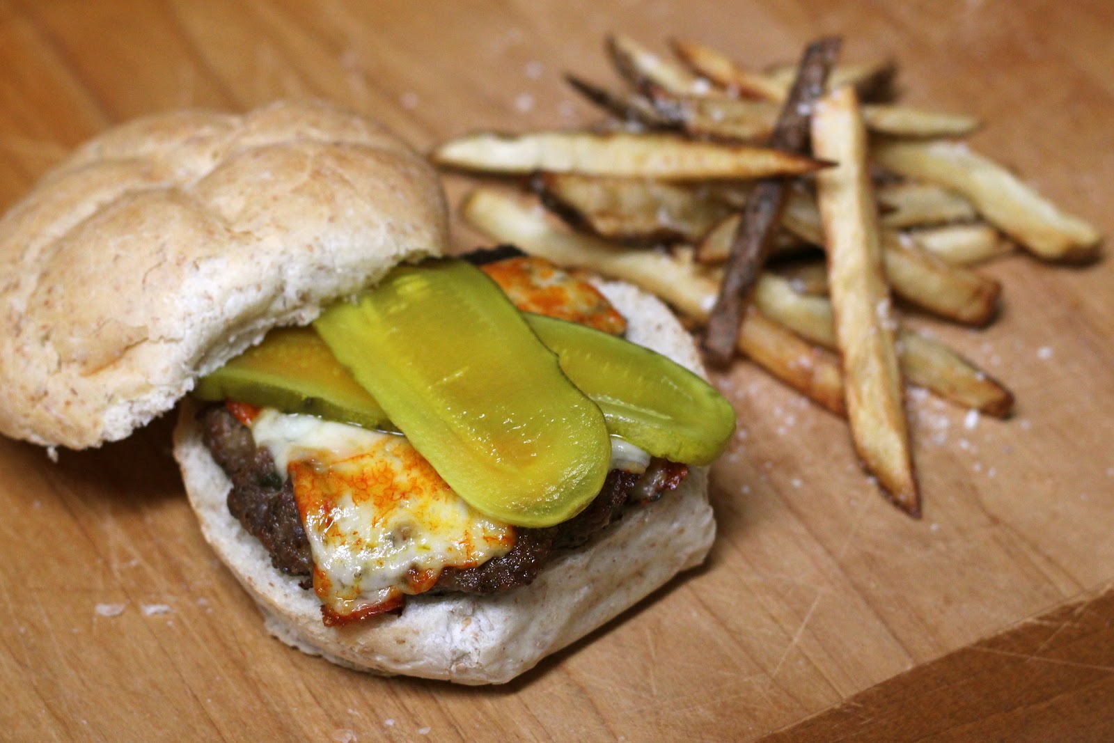 A juicy venison cheeseburger with melted cheddar on a toasted bun, sitting on a plate with pickles and a small pile of fries on a wooden table in warm natural light