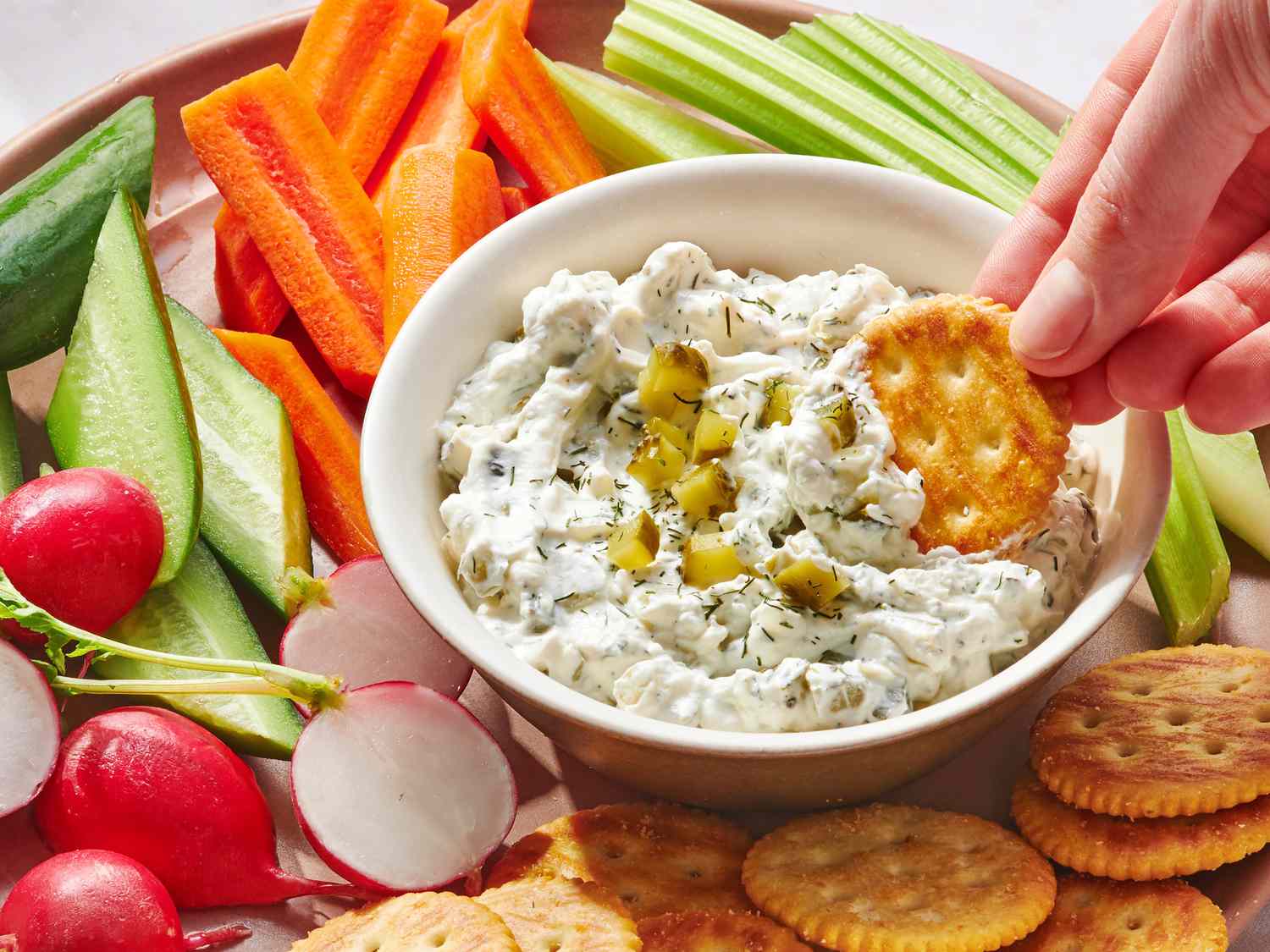 A kitchen counter with a jar of dill pickles, blocks of cream cheese, sour cream, fresh dill, garlic, and lemon arranged as mise en place in warm natural light