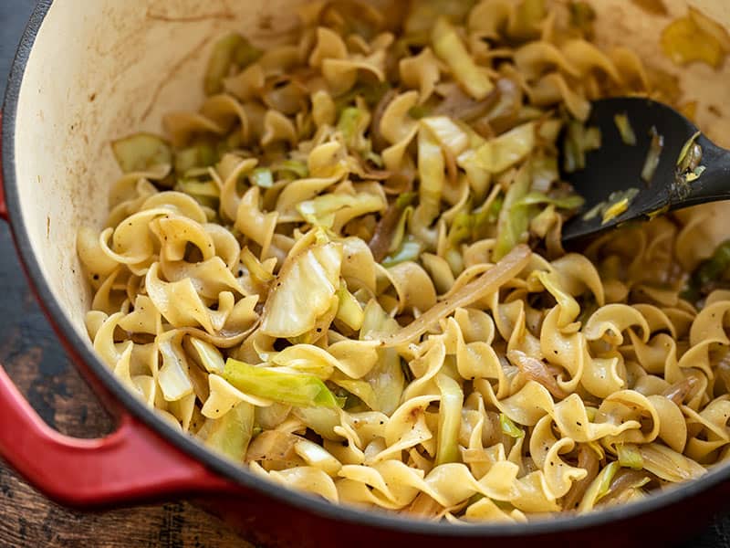 A kitchen counter with a whole green cabbage, a bag of egg noodles, a yellow onion, a stick of butter, salt, and a pepper grinder arranged for cooking