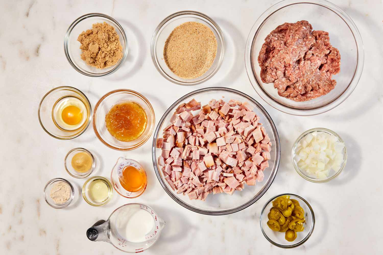 A kitchen counter with bowls of ground ham and ground pork, crushed crackers, eggs, milk, diced onion, ketchup, brown sugar, vinegar, and mustard set out for ham loaf prep in warm natural light