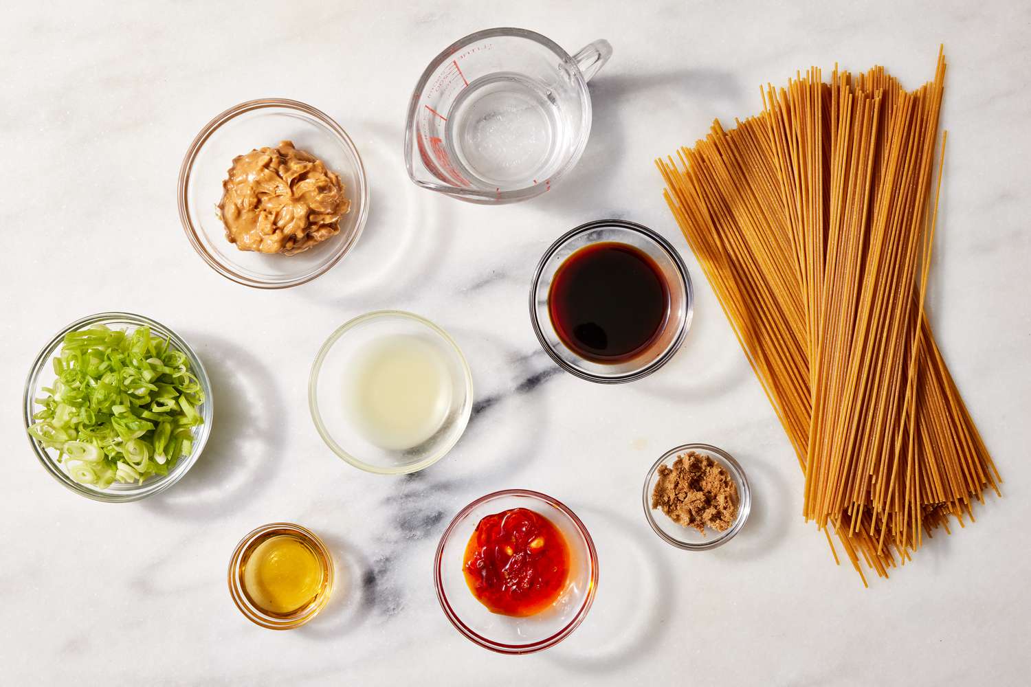 A kitchen counter with dry rice noodles, peanut butter, soy sauce, lime, garlic, ginger, honey, chili paste, and fresh vegetables laid out for cooking