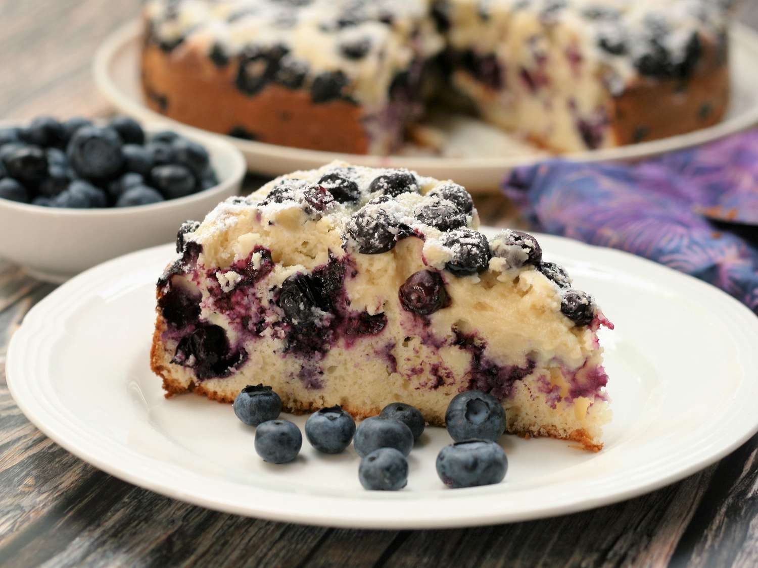 A kitchen counter with lemons, a microplane, fresh blueberries, a bowl of gluten free flour blend, sugar, and baking powder arranged for baking
