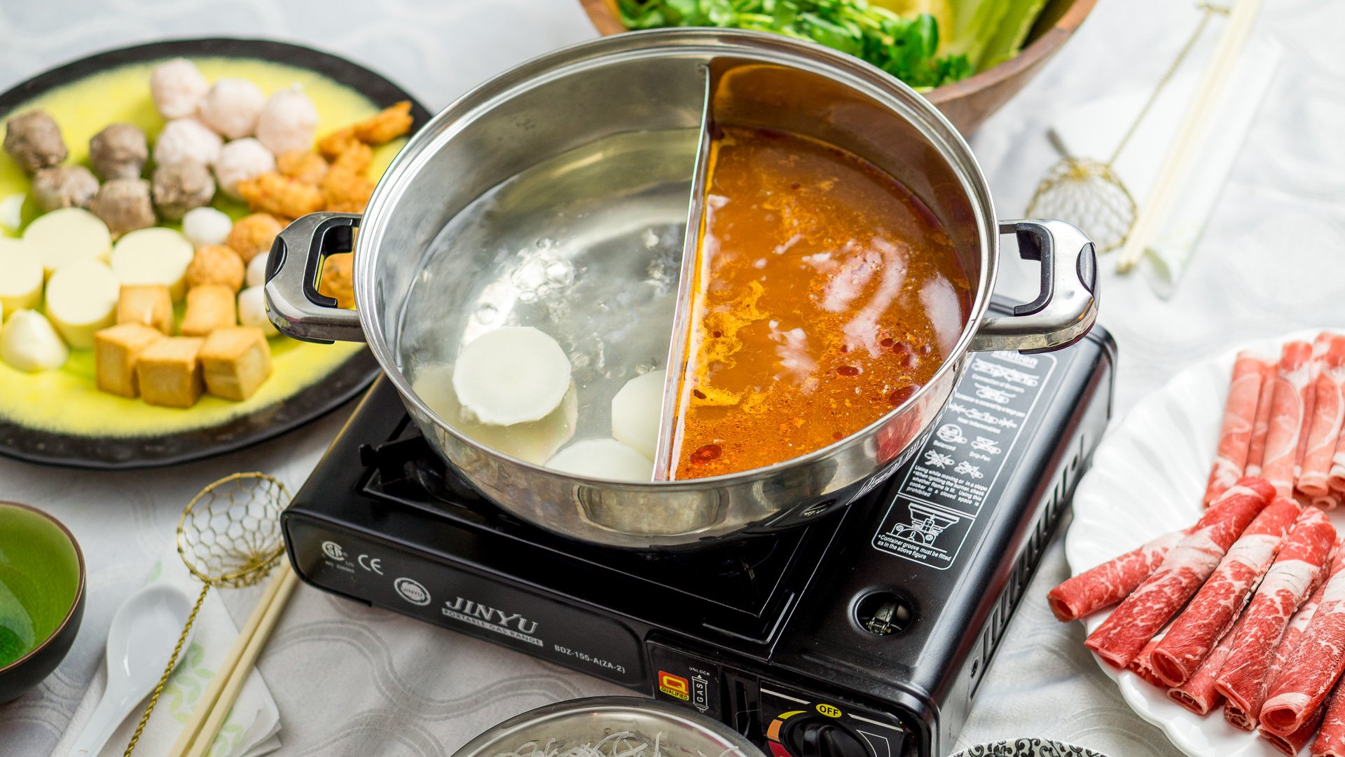 A kitchen counter with neatly arranged shabu shabu ingredients including kombu, napa cabbage, shungiku or spinach, enoki mushrooms, shiitake mushrooms, tofu, thinly sliced beef, udon noodles, and small bowls of condiments