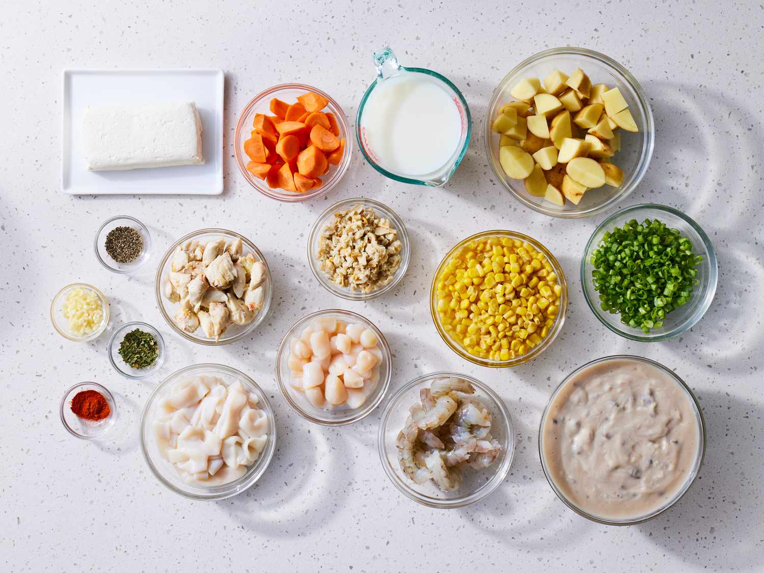 A kitchen counter with raw shrimp, white fish pieces, bacon, potatoes, onion, celery, garlic, corn, thyme, cream, broth, and clam juice arranged in small bowls