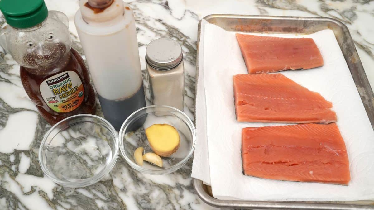 A kitchen counter with small bowls of soy sauce, honey, lemon juice, minced garlic, grated ginger, and a spoonful of cornstarch next to raw salmon fillets